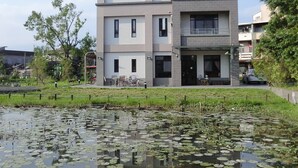 A multi-story residential building with a flat roof, large windows, and a balcony. In front of the building, there is a grassy area with a pond covered in lily pads. - Love Play House (Luodong)