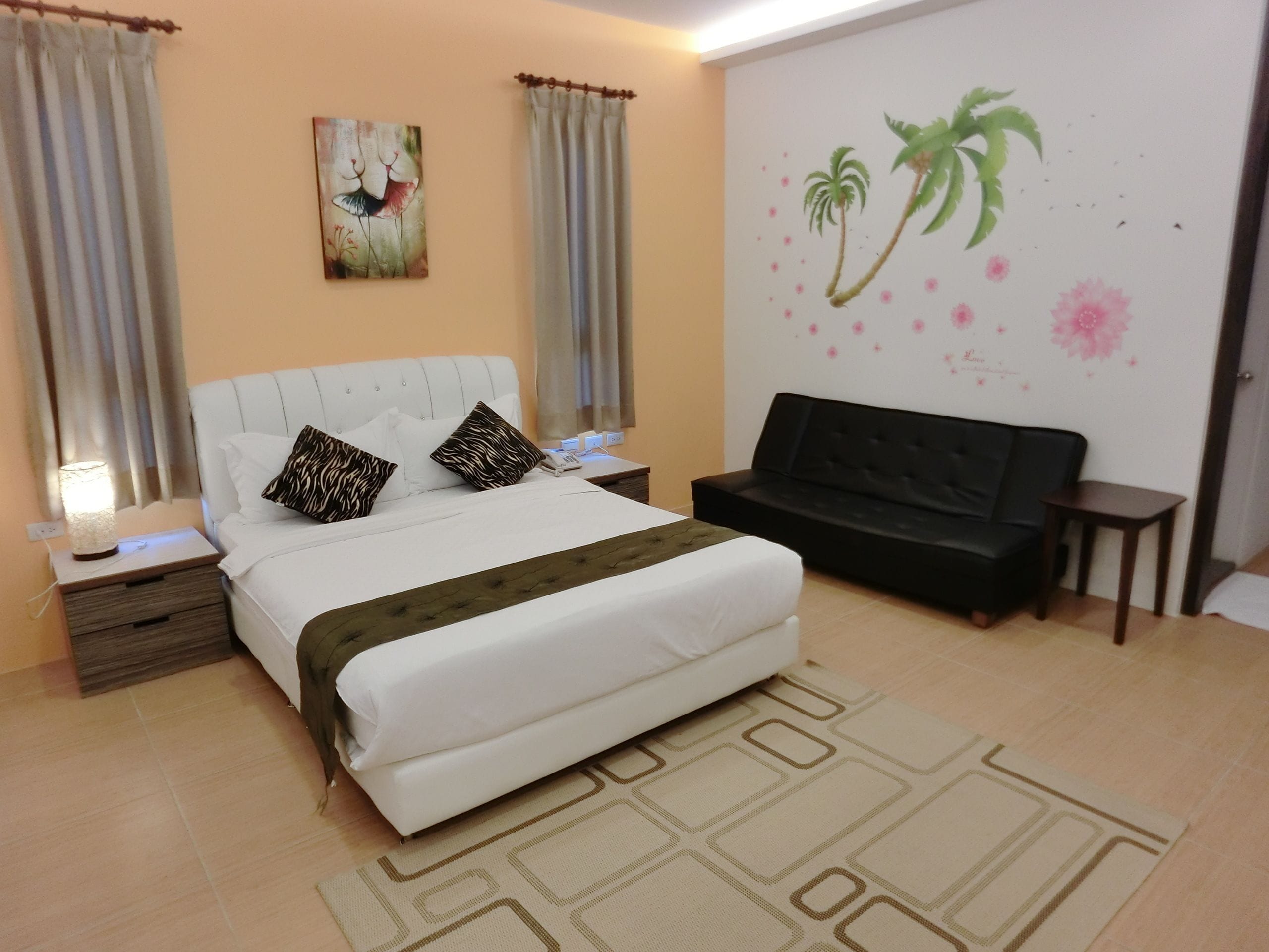 Un dormitorio con cama, mesita de noche, sofá negro, pared con un mural floral y ventana con cortinas.