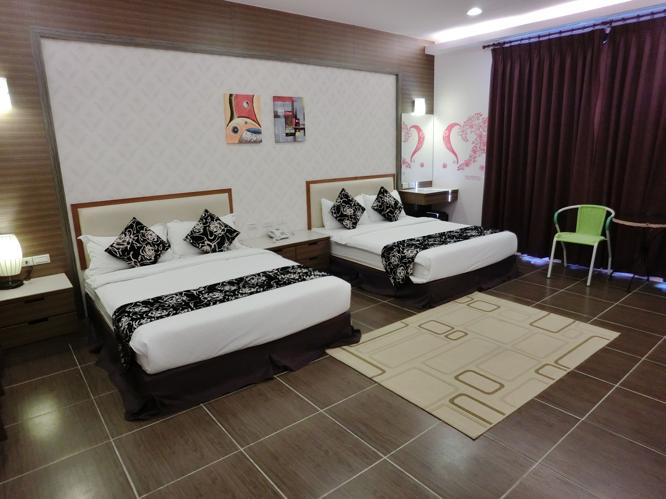 A hotel room with two beds, a nightstand with a lamp, a mirror, a green chair, and artwork on the wall.