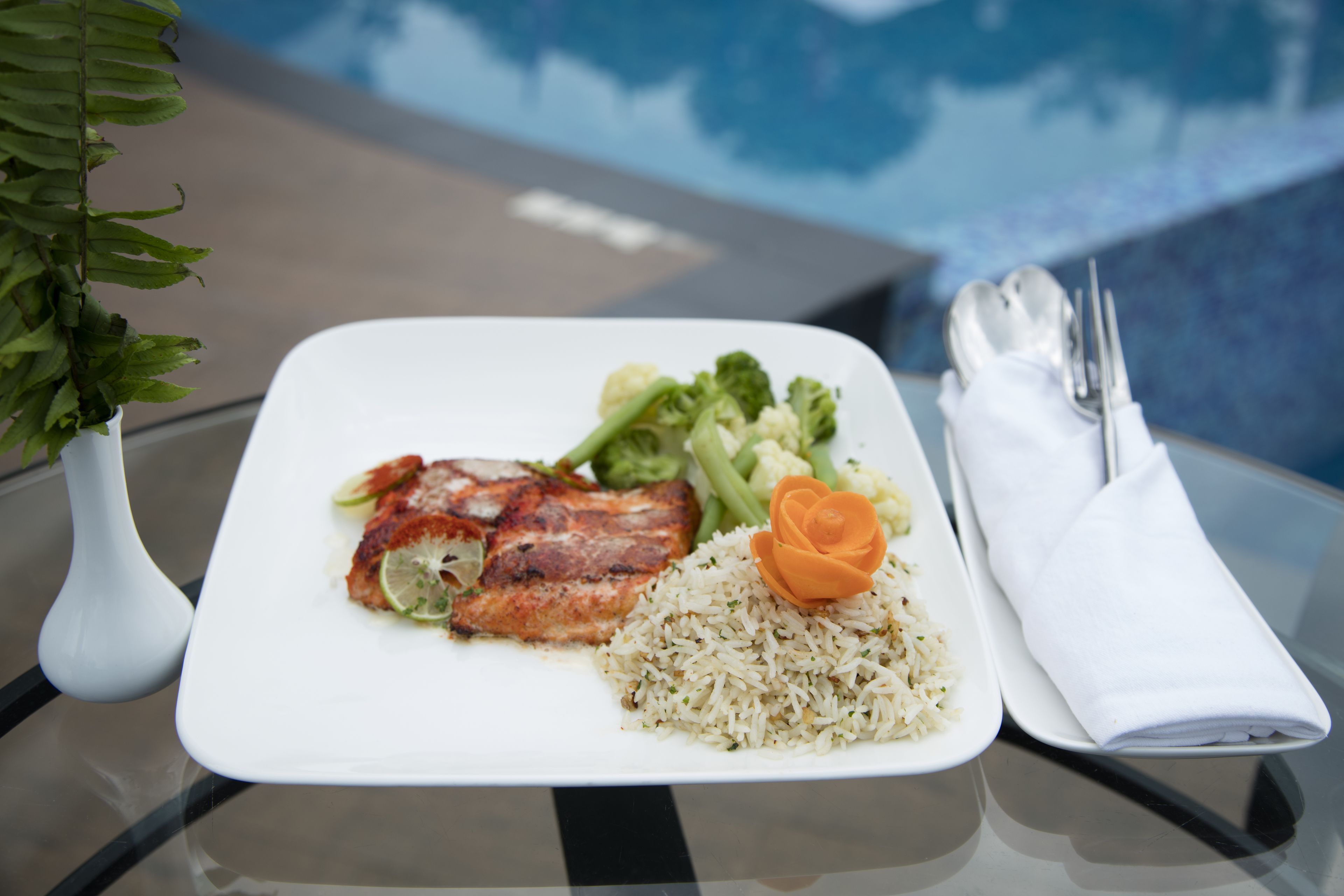 A plate of food with rice, vegetables, and meat, accompanied by a napkin and utensils on a glass table near a pool.