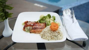 A plate of food with rice, vegetables, and meat, accompanied by a napkin and utensils on a glass table near a pool.