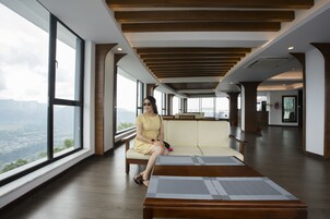 Un amplio vestíbulo de hotel con un ventanal que ofrece vistas de la ciudad, una zona de estar con sofá y una mesa, y un techo de madera.