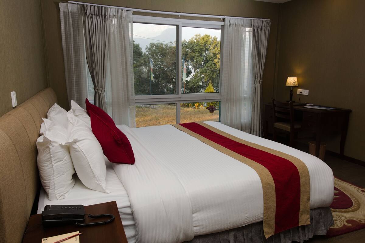 A hotel room with a large bed, a desk with a lamp, and a view of trees through the window.