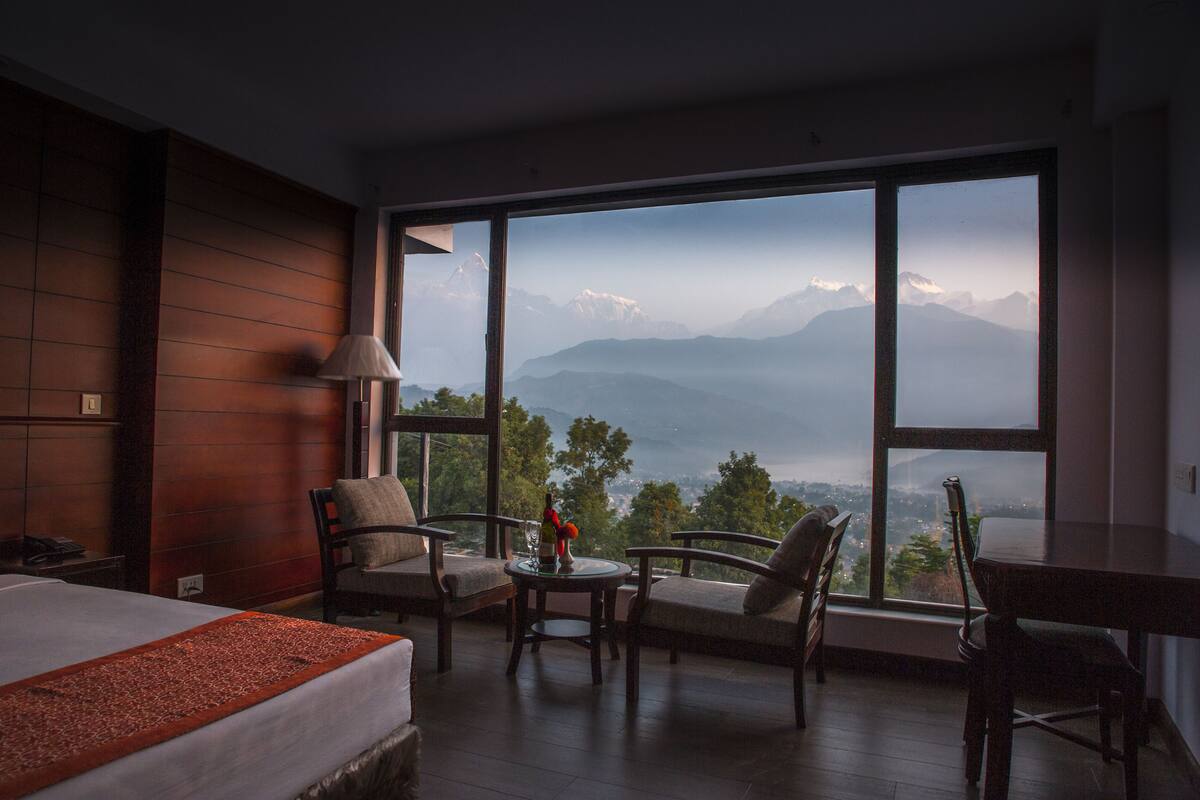 A hotel room with a large window, a bed, a table with a vase, and two chairs.