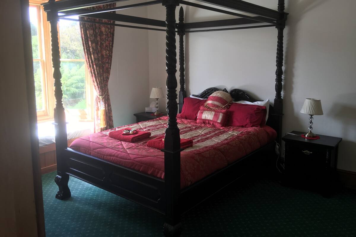 A four-poster bed with a red bedspread and pillows, a nightstand with a lamp, and a window with curtains.