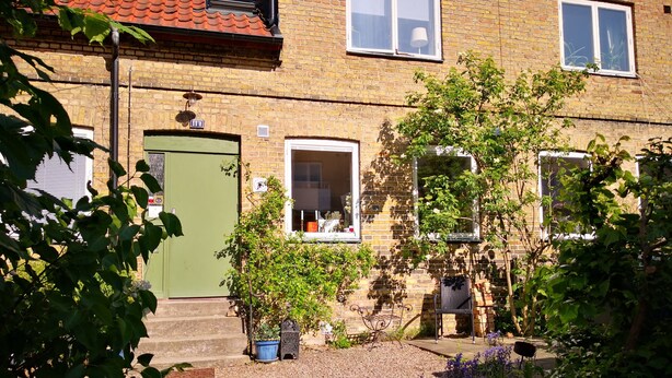 A two-story brick house with a green door, multiple windows, and a small garden area with gravel, potted plants, and a metal bench. - Explorers Club (Lund)