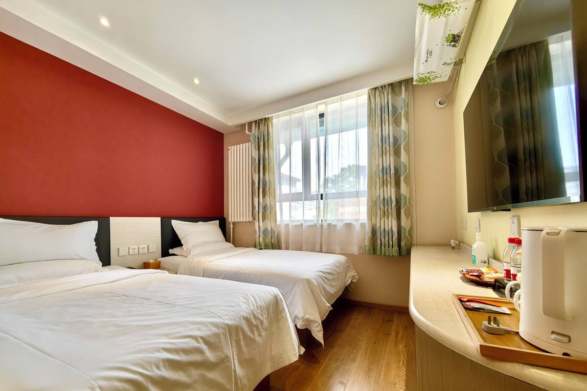 A hotel room with two beds, a wooden floor, a red accent wall, a large window with sheer curtains, and a flat-screen TV mounted on the wall.