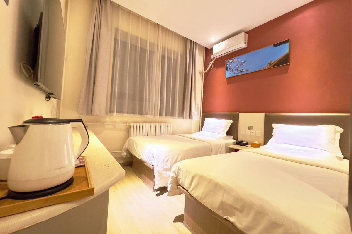A hotel room with two beds, a coffee maker, a bedside table, a window with curtains, an air conditioning unit, and a painting on the wall.