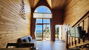 A wooden cabin interior with a sofa, a table with a lace tablecloth, a TV, and a large arched window offering a view of the sea. - Old Man and Sea Pension (Seogwipo)