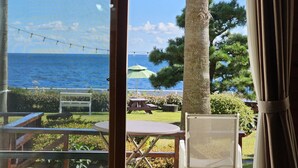 A view from a room with a glass door to a patio area. The patio has a table, chairs, and an umbrella. There is a palm tree and a view of the ocean. - Old Man and Sea Pension (Seogwipo)