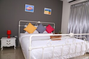 A neatly made bed with a white metal frame, a bedside table with a red lamp, and two wall-mounted pictures. - Apartment Khunpa (Koh Samui)