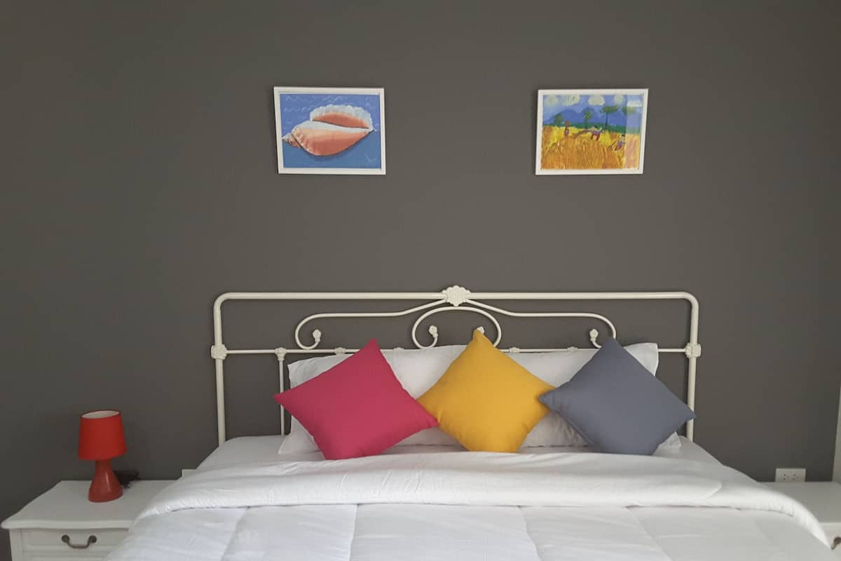 A neatly made bed with white bedding and three colorful pillows. There is a white nightstand with a red lamp beside the bed. Two framed artworks are hung on the wall above the bed.