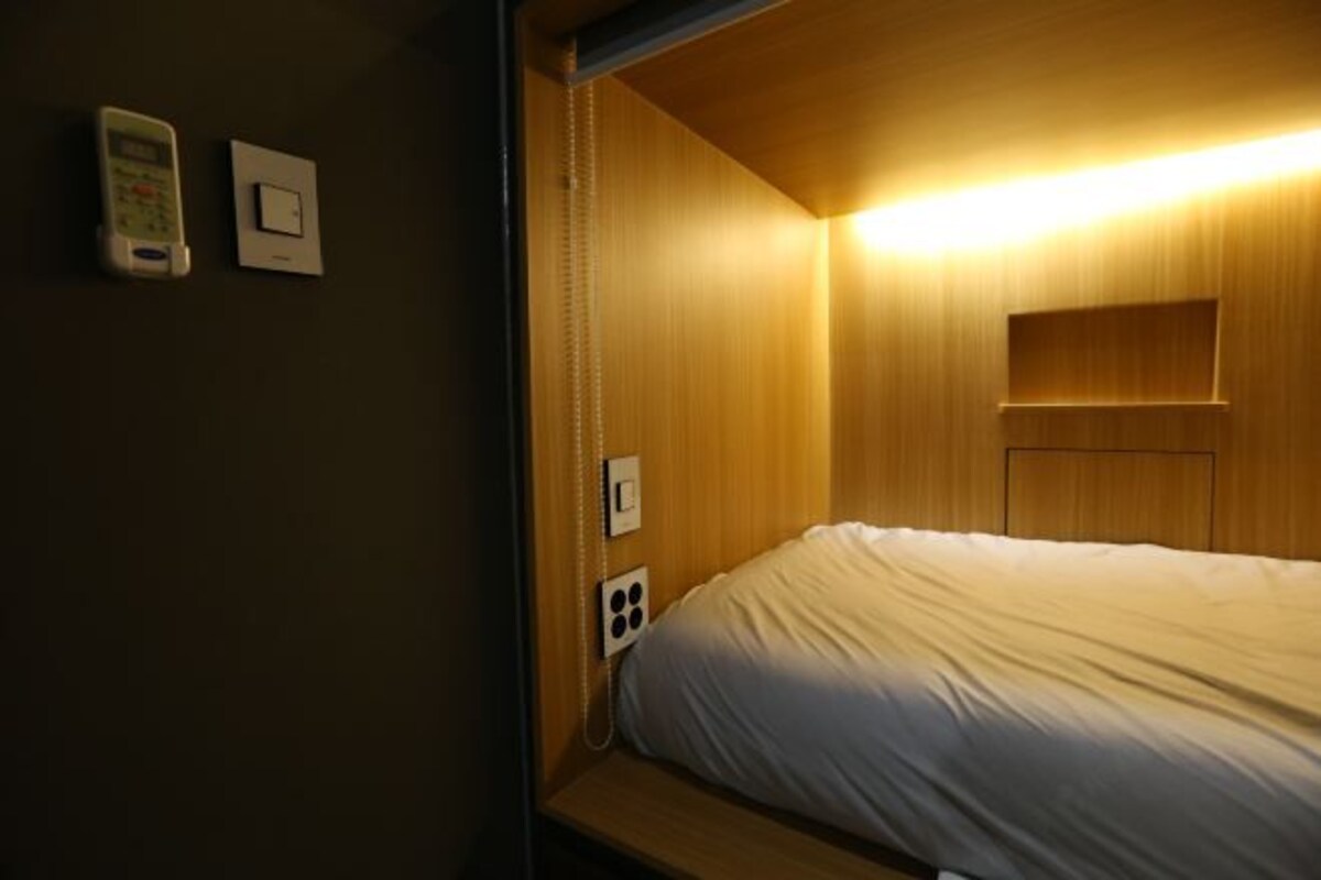 Une petite pièce faiblement éclairée, avec un lit simple, une tête de lit en bois et un téléphone fixé au mur.