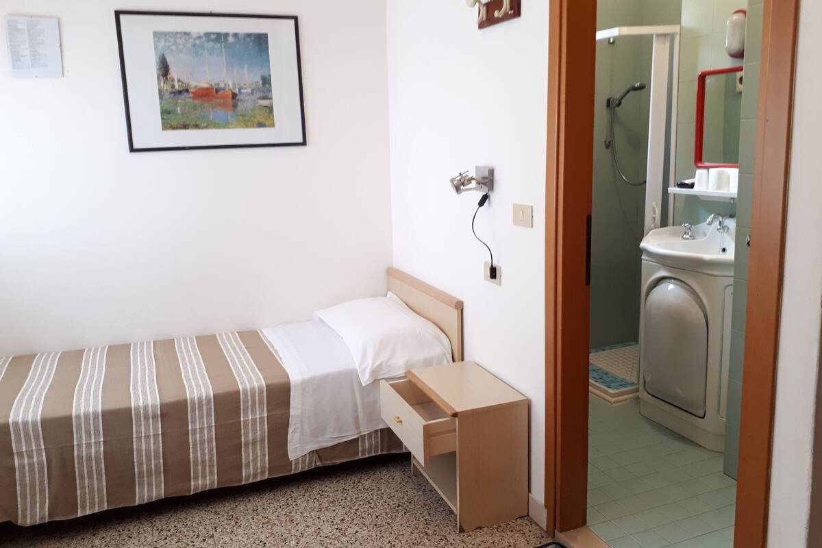 Una habitación pequeña con cama individual, un escritorio de madera, un cuadro en la pared y un baño visible con lavadora.