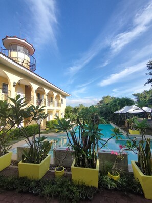 A pool area with a building in the background, surrounded by potted plants and umbrellas. - Villa Del Faro (Calatagan)