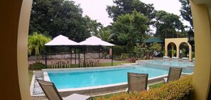 A swimming pool with lounge chairs and umbrellas, surrounded by greenery and a building with arches. - Villa Del Faro (Calatagan)