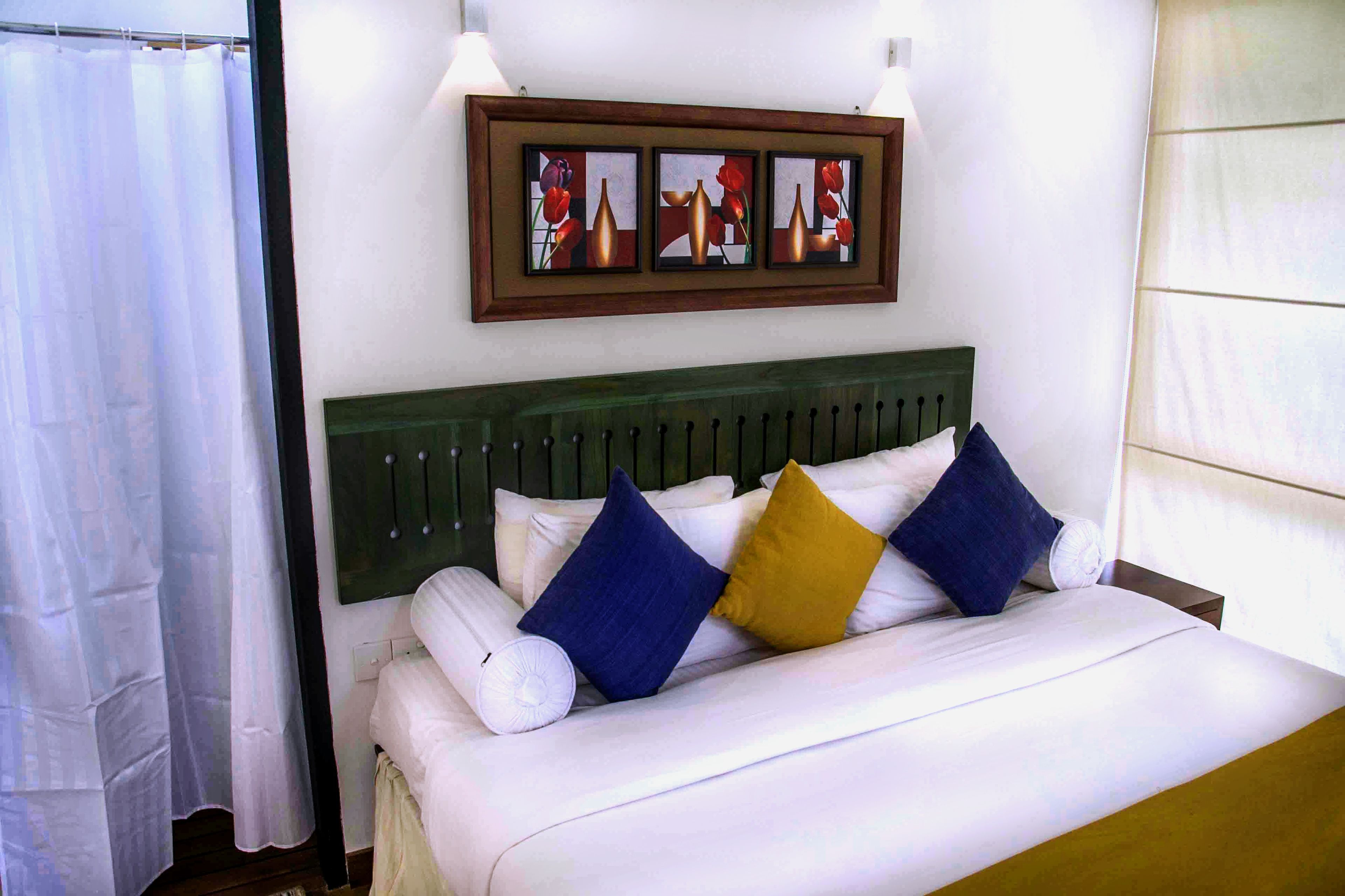 Una cama bien tendida con ropa blanca y tres almohadas en azul, amarillo y blanco.