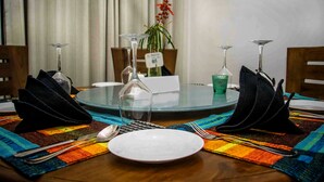 A round dining table with a glass top, surrounded by chairs, a vase with a plant, and a placemat. - Hotel Yo (Kandy)