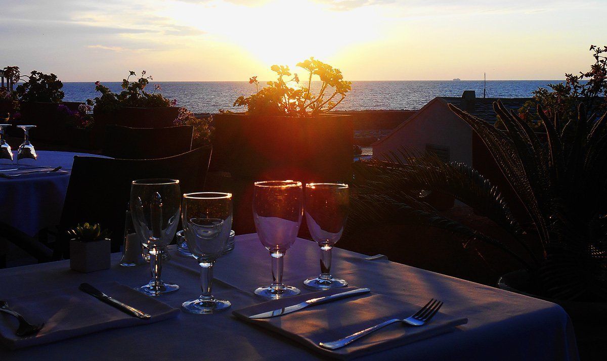 A table set for two with wine glasses, a water glass, and cutlery, overlooking a sunset over the sea.