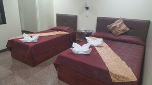 A hotel room with two single beds, each with a red bedspread and white towels. There is a nightstand with a remote control and a lamp.