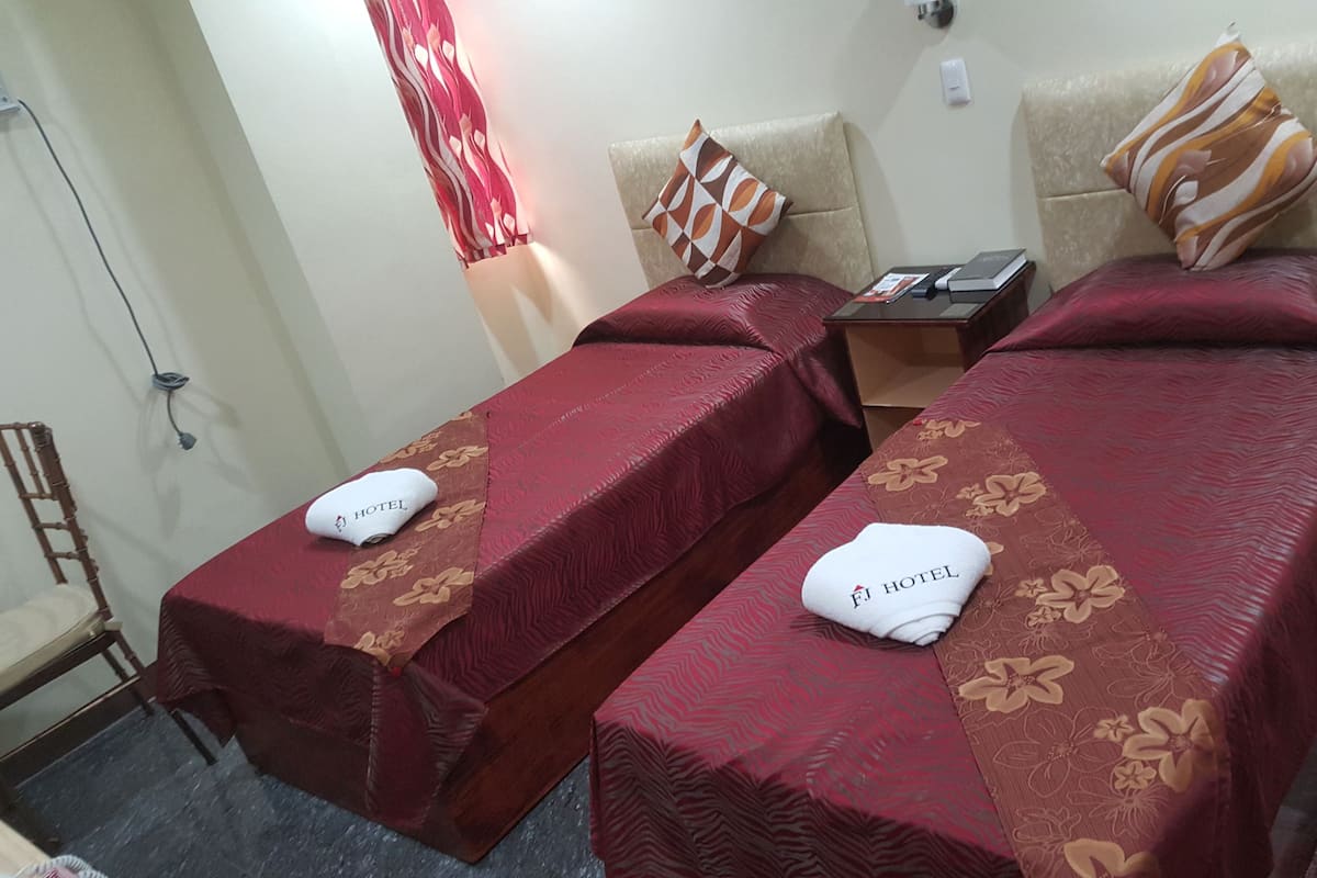 Two single beds with red and brown floral bedspreads, each with a white towel labeled "FJ HOTEL" and a pillow with a geometric pattern.