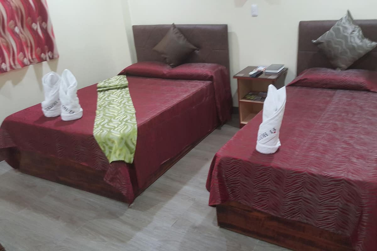 Two single beds with red bedding and pillows, a small wooden nightstand with a book and a remote control, and a wall-mounted light switch.