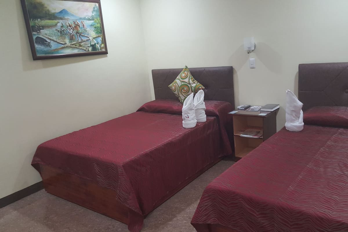 A hotel room with two single beds, each with a red bedspread and a towel. There is a painting on the wall, a nightstand with a lamp, and a small table with a book and a remote control.