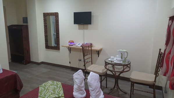 A hotel room with a bed, a small table with a kettle, a chair, a desk with a phone and lamp, and a TV on the wall. - FJ Manila Hotel (Daraga)