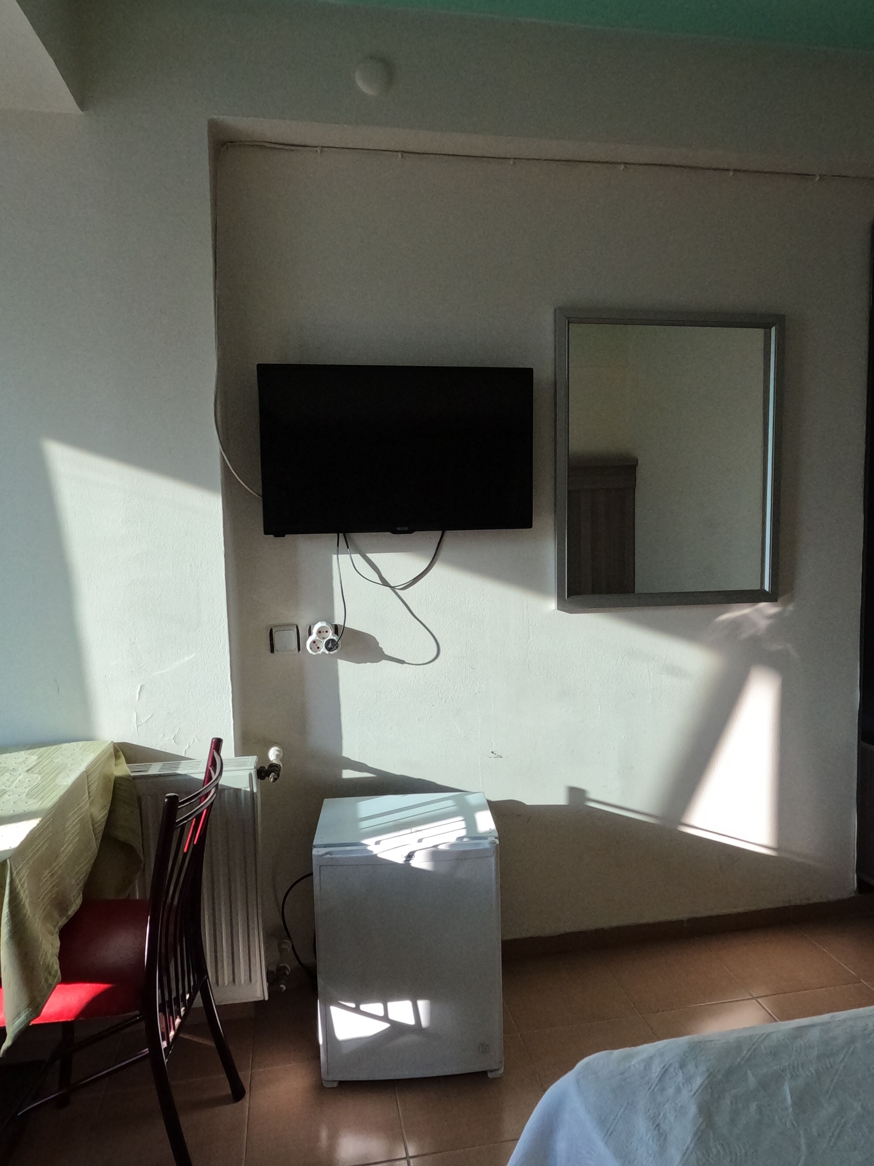 A room with a flat-screen TV mounted on the wall, a small refrigerator, a chair, and a bed.