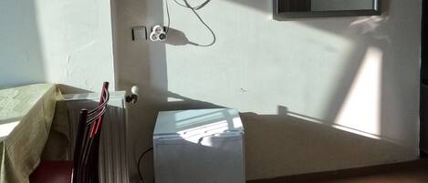 A room with a flat-screen TV mounted on the wall, a small refrigerator, a chair, and a bed.