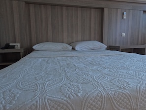 A neatly made bed with a patterned bedspread, a wooden headboard, and a bedside table with a phone and a lamp. - Cihanay Hotel (Antalya)