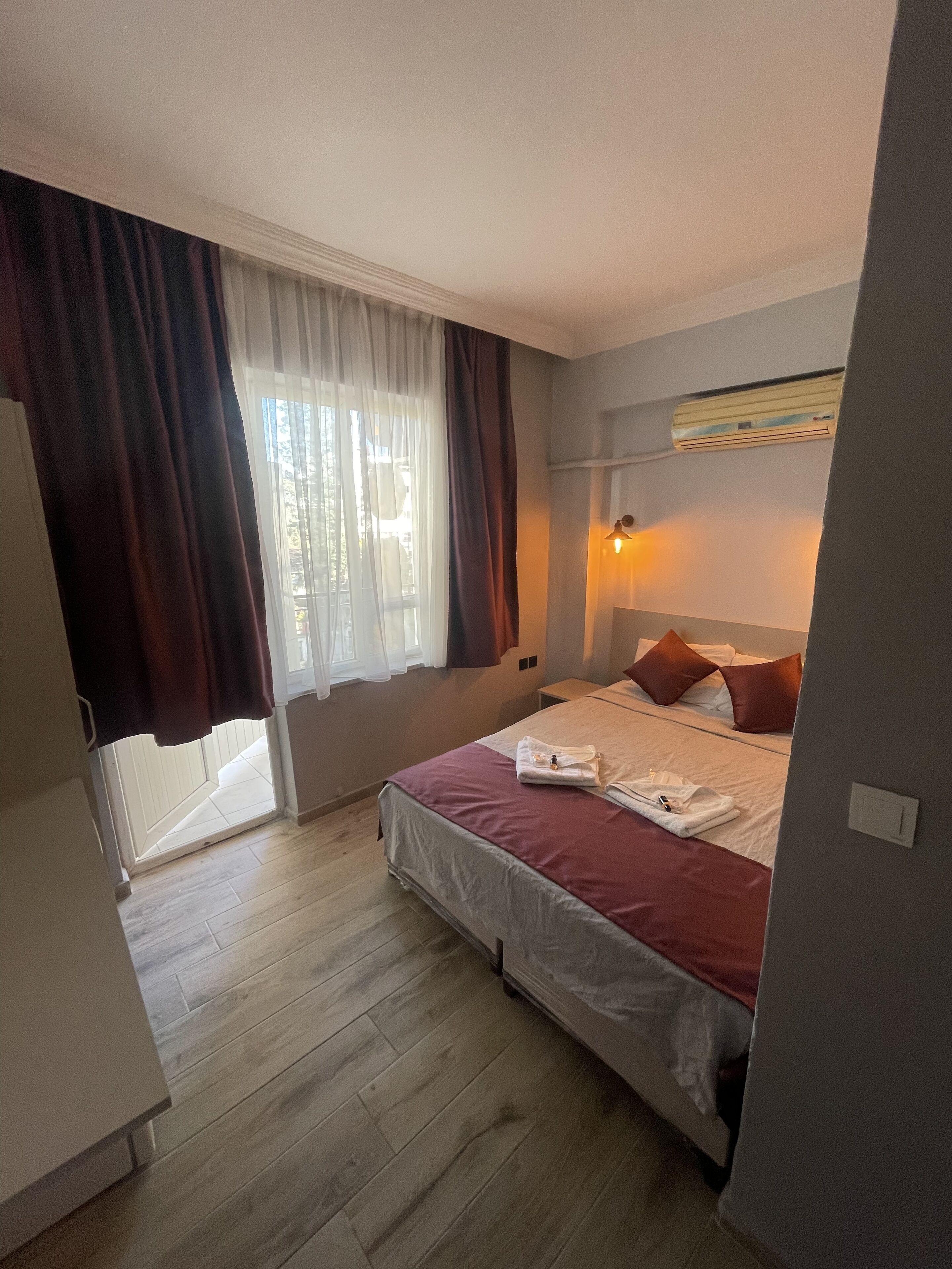 A hotel room with a bed, a nightstand, a window with curtains, an air conditioning unit, and a wooden floor.