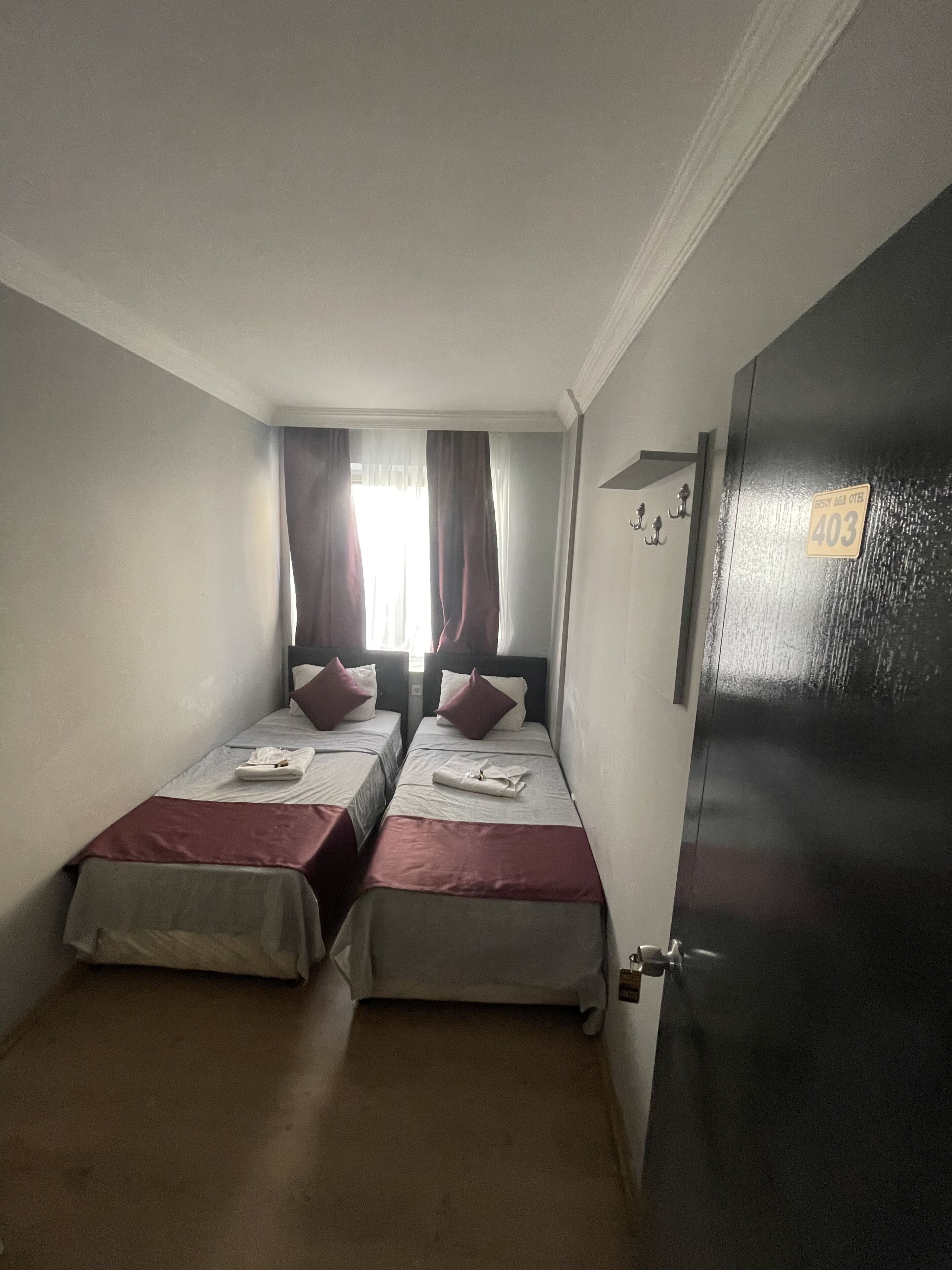 Una habitación pequeña y tenuemente iluminada con dos camas individuales, cada una con una colcha roja y almohadas blancas. Una ventana con cortinas permite la entrada de luz natural. Se ve una puerta con el número a la derecha.