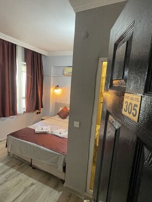 Una camera d'albergo con un letto, una finestra con tende, una lampada a parete e una porta con il simbolo del numero.