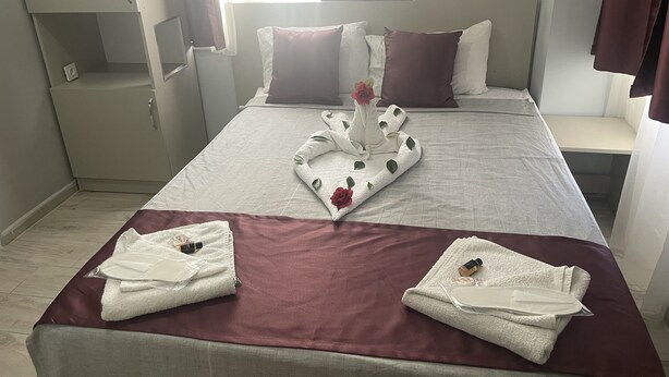 A neatly made bed with a towel arrangement, a bedside table, and a nightstand. - Ersoy Aga Otel (Antalya)