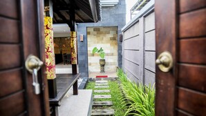 Uma passagem estreita que leva à entrada de um edifício, com porta de madeira, um pequeno jardim com planta em vaso e chão de azulejos.