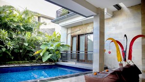 Uma piscina com deck ladrilhado, cercada por vegetação exuberante e um edifício moderno com varanda.