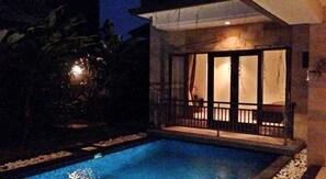 A swimming pool with a wooden deck and a glass door leading to a covered area. - Legong Kirana Villas (Legian)