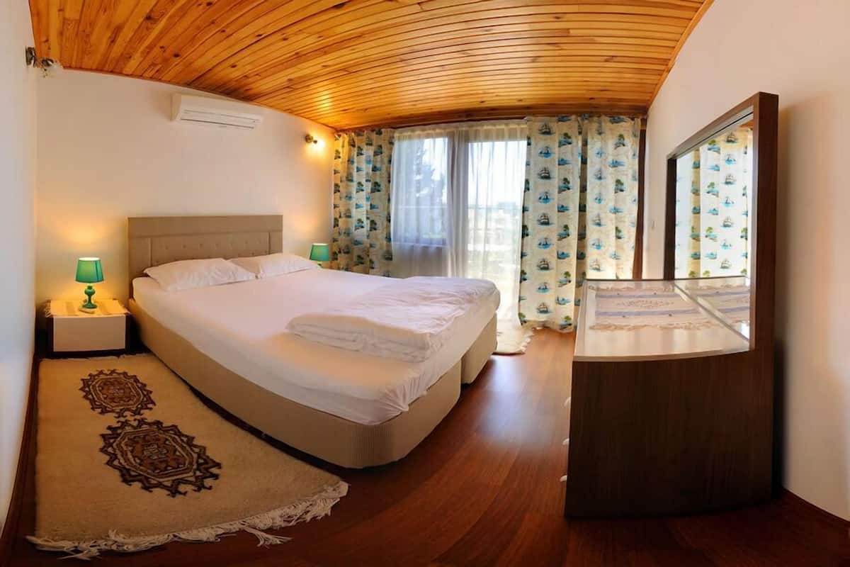 Een slaapkamer met een houten plafond, een bed met witte lakens, een nachtkastje met een lamp en een raam met patroon gordijnen.