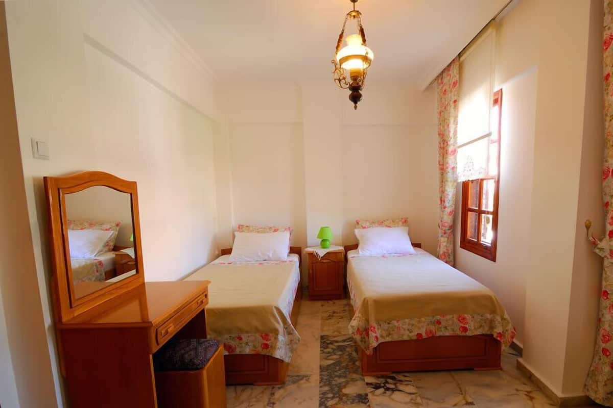 Een slaapkamer met twee enkele bedden, een houten dressoir, een spiegel, een kroonluchter en een raam met bloemen gordijnen.
