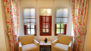 Una habitación con dos sillones color beige enfrentados, una mesita con un jarrón de flores y una ventana con contraventanas de madera y un cristal decorativo.