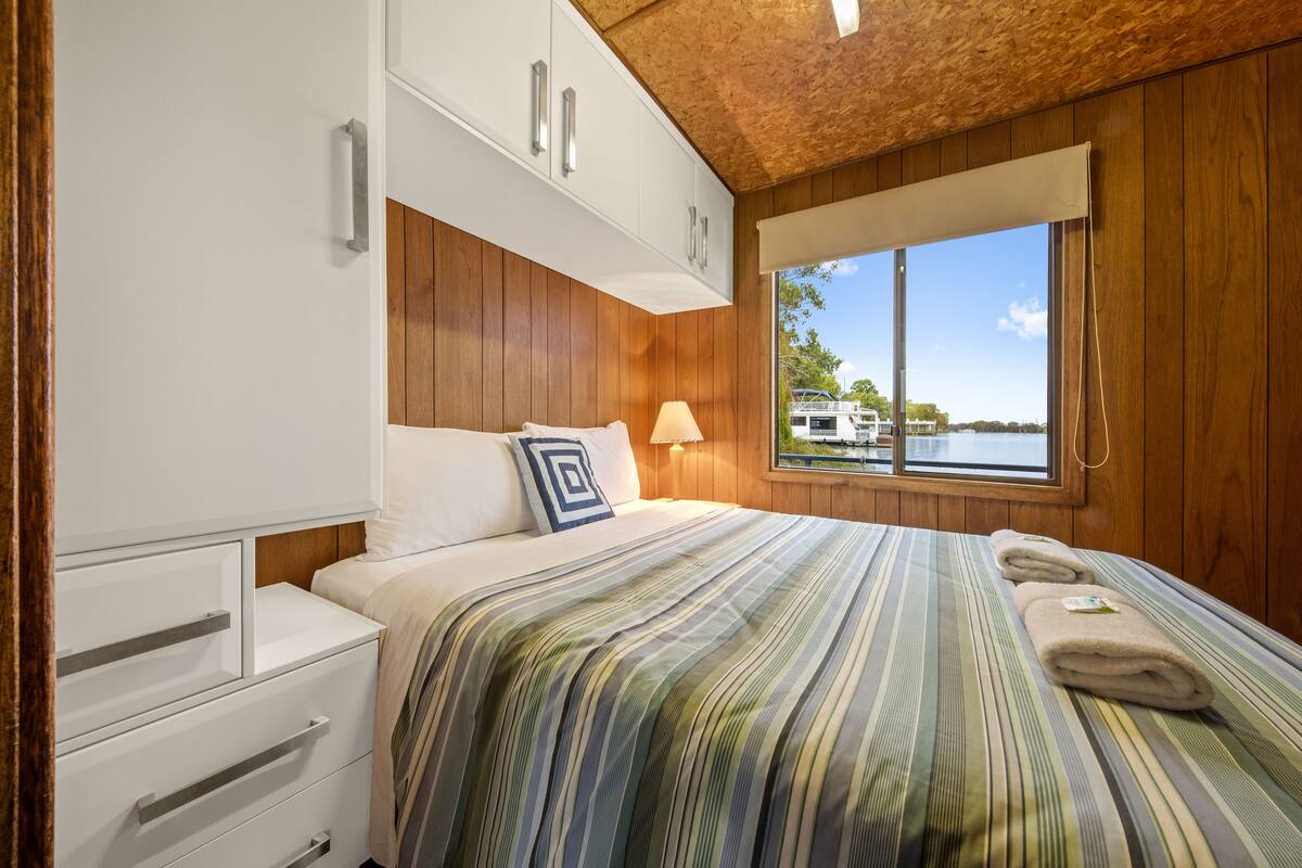 Un dormitorio con techo de madera, una cama con ropa de cama a rayas, una mesita de noche, una ventana con vista a un barco y armarios empotrados blancos.