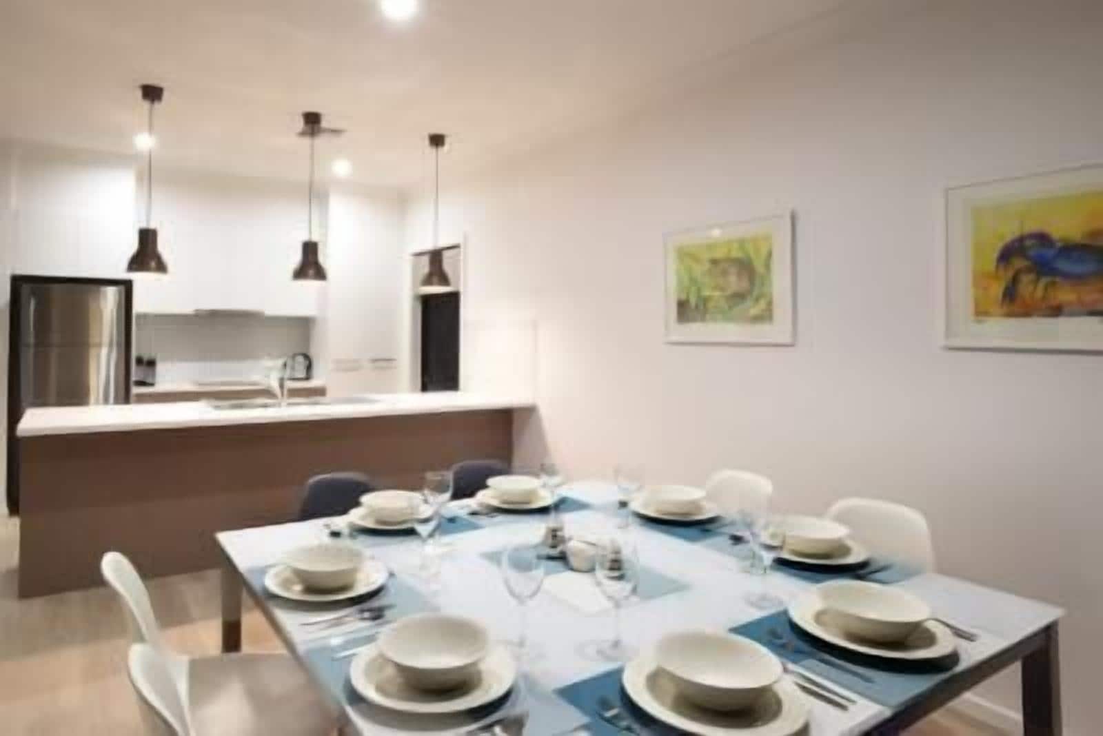 Un espace repas moderne avec une table dressée pour six personnes, une cuisine en arrière-plan et deux œuvres d’art encadrées accrochées au mur.