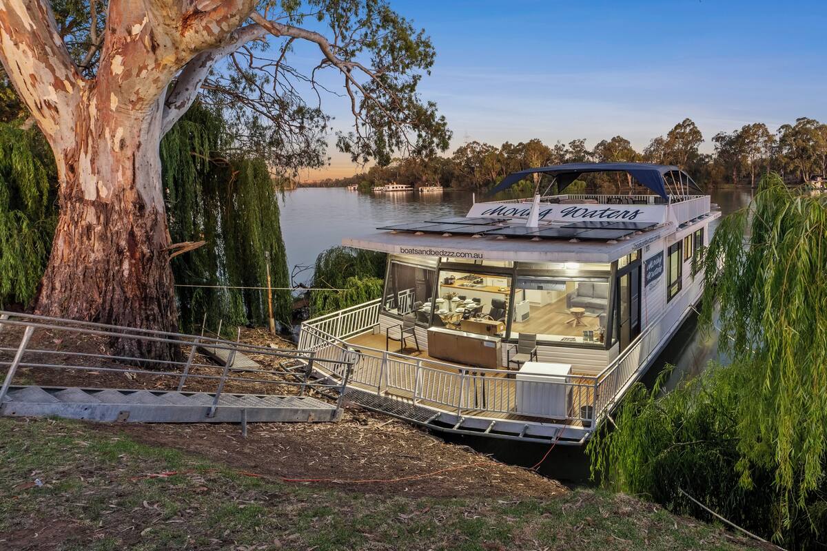 Un barco de paseo fluvial con cubierta techada y comedor, atracado cerca de un árbol grande y agua.