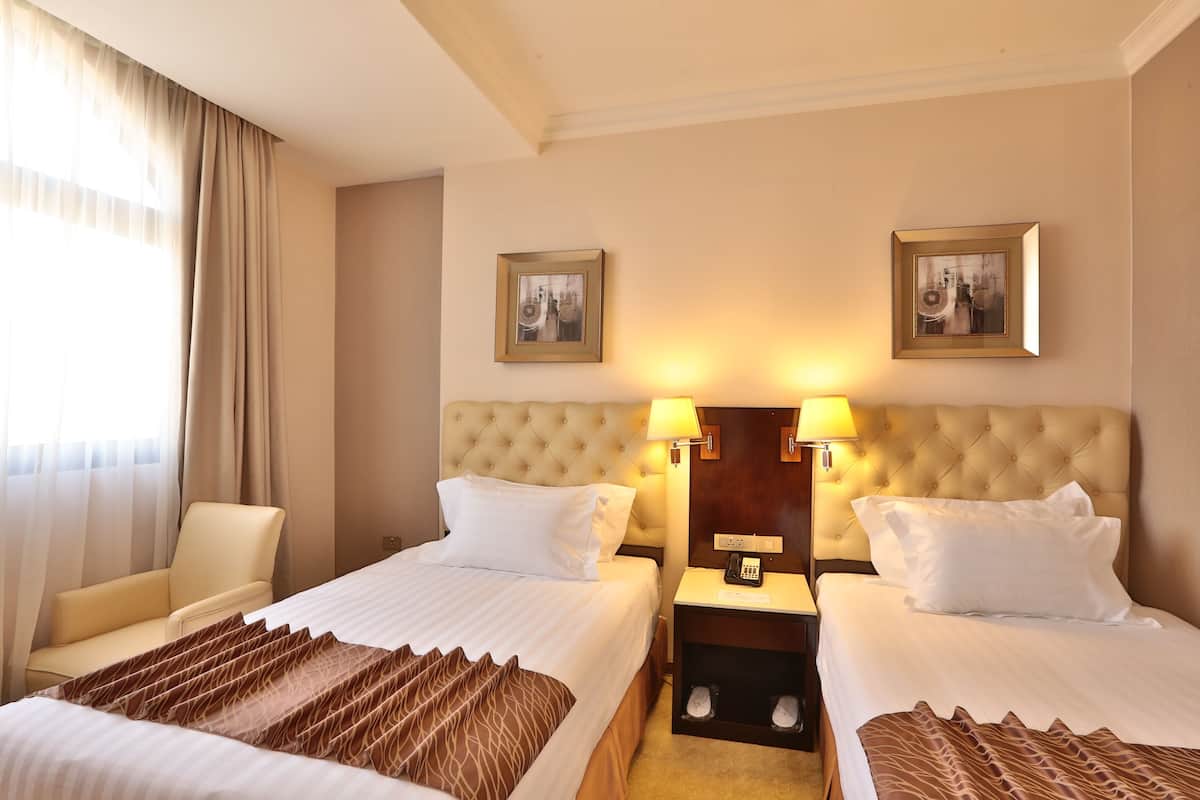 A hotel room with two beds, a chair, a nightstand with a lamp, and two framed pictures on the wall.
