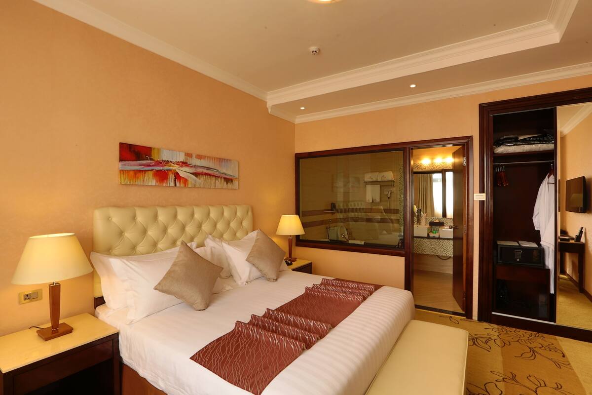 A hotel room with a large bed, two bedside tables, a mirror, a painting on the wall, and a view into the bathroom.