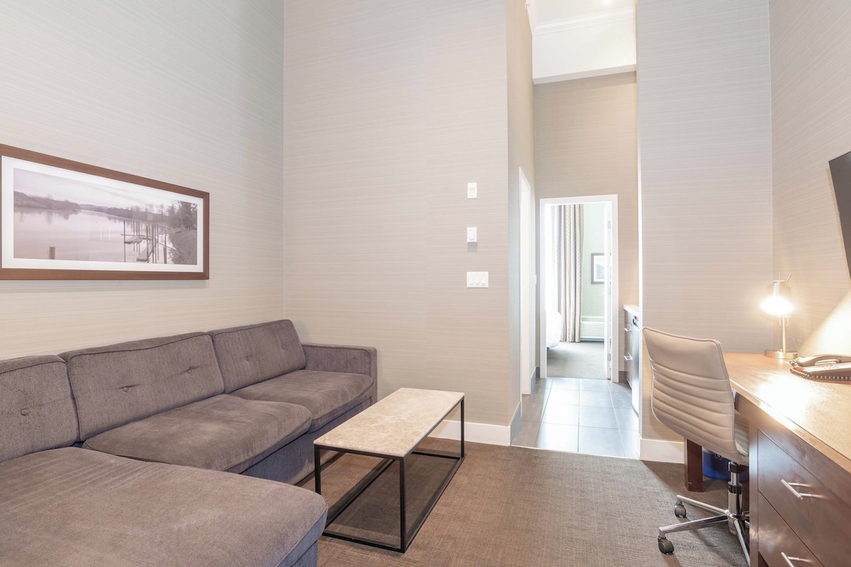 A modern living room with a gray sofa, a small table, and a desk with a chair. There is a framed picture on the wall and a hallway leading to another room.