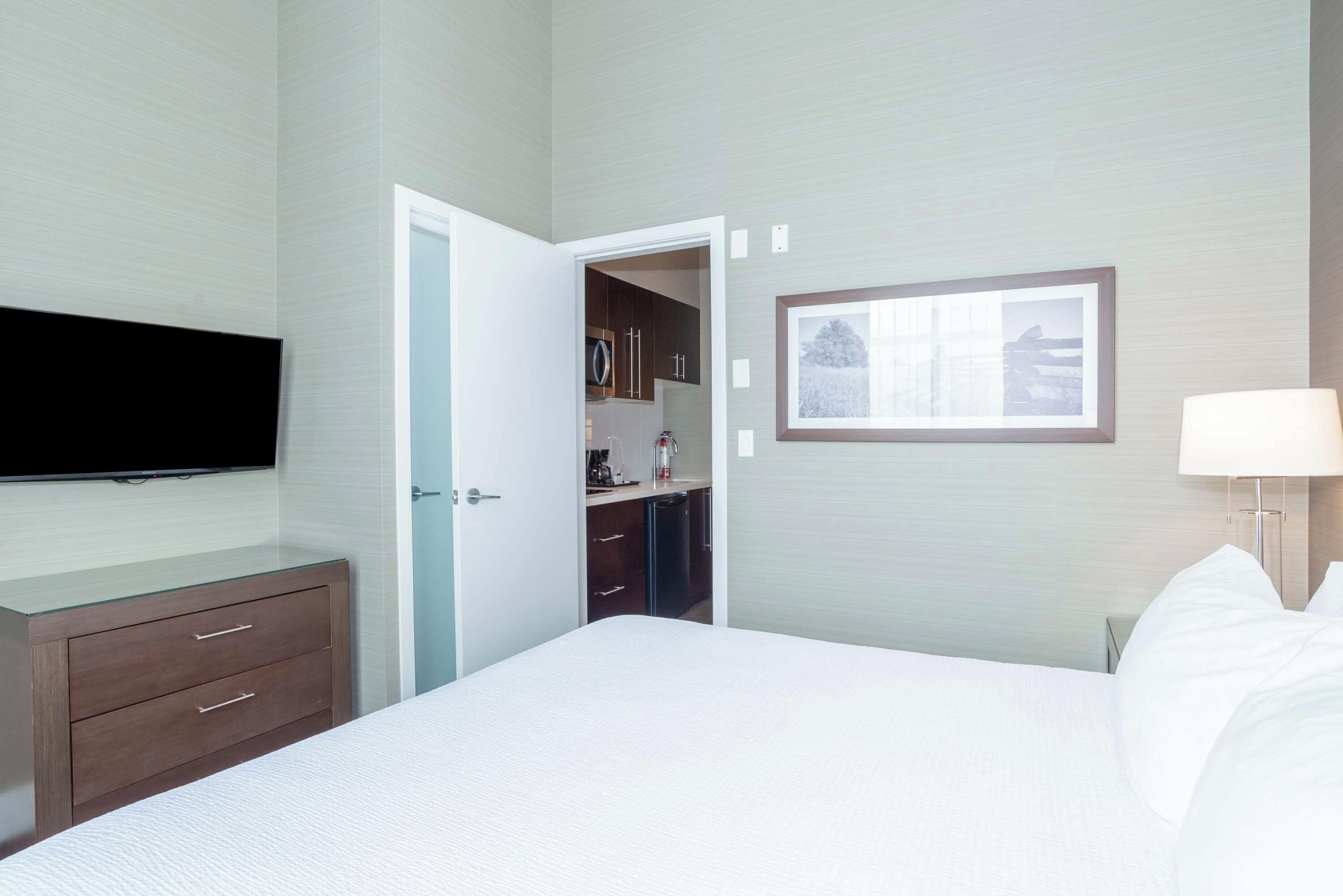 A hotel room with a bed, a TV mounted on the wall, a bedside table with a lamp, and a door leading to a bathroom.
