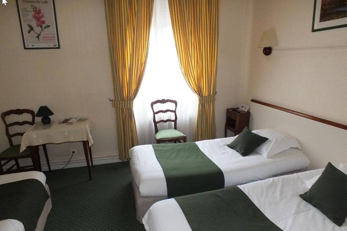 Habitación de hotel con dos camas, una mesita con lámpara, una silla y una ventana con cortinas.