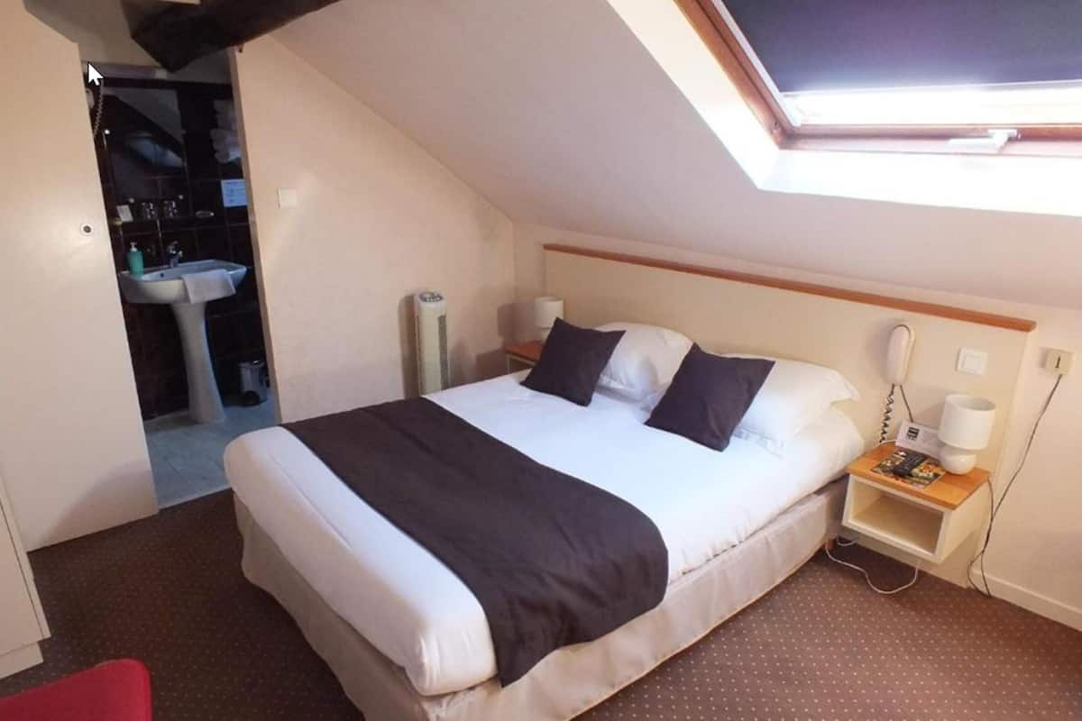 Habitación de hotel con una cama individual, una mesita de noche con lámpara, un teléfono y un estante pequeño. Hay una ventana sobre la cama que proporciona luz natural.